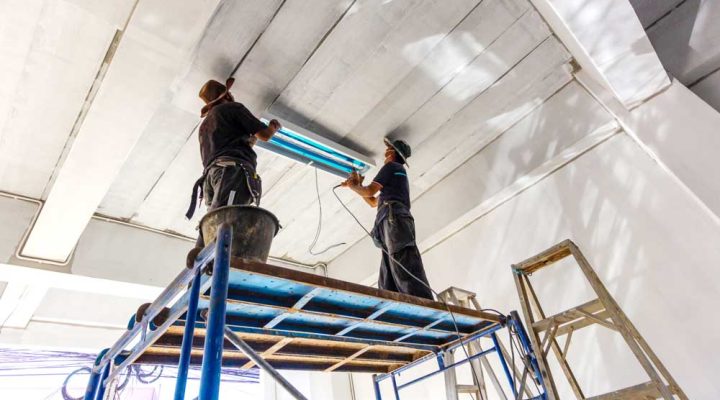 Two workers on scaffolding install fluorescent lighting fixtures in a high-ceilinged commercial building.